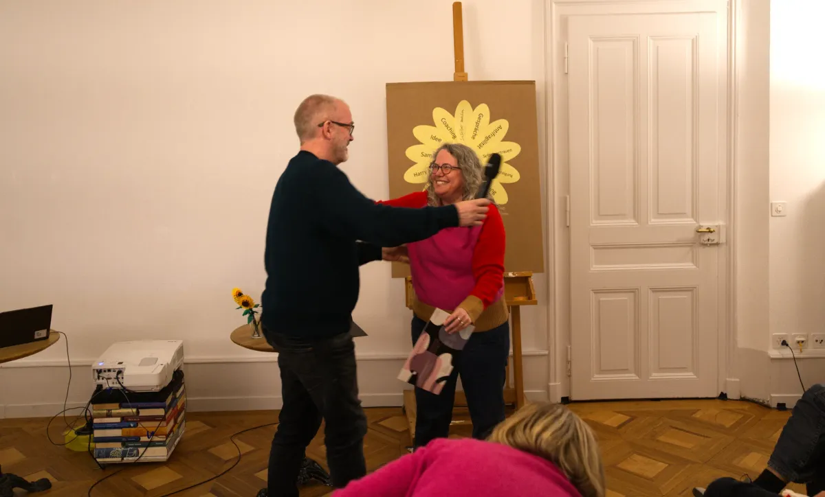 Abschlussfeier der Colearning Akademie: Ein Lerncoach umarmt eine Absolventin vor einer Tafel mit einer gelben Blume aus Begriffen
