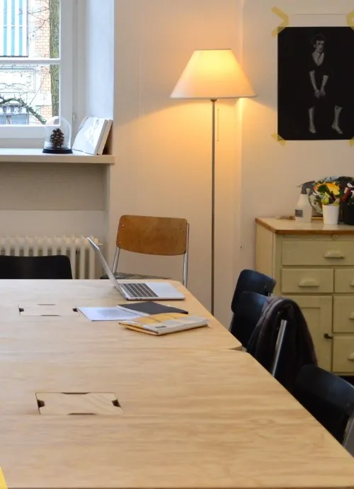 Lernraum am Colearning-Tag: langer Holztisch mit Laptop und Notizen, Stühle, Stehlampe und eine Kommode mit Blumen